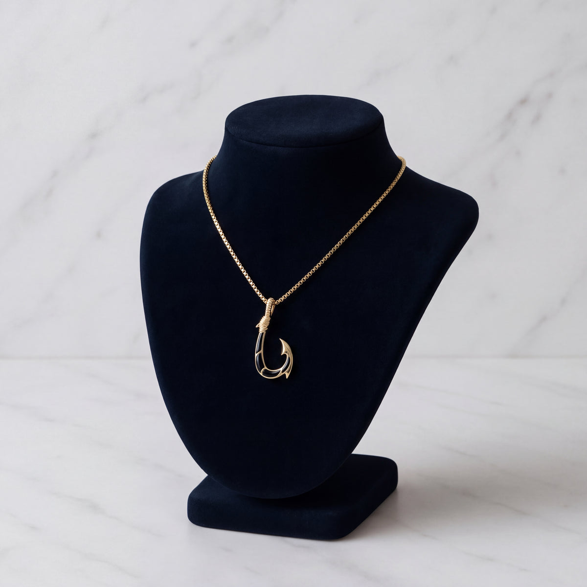 A gold fish hook pendant with a dark inlay, on a navy velvet bust over marble.