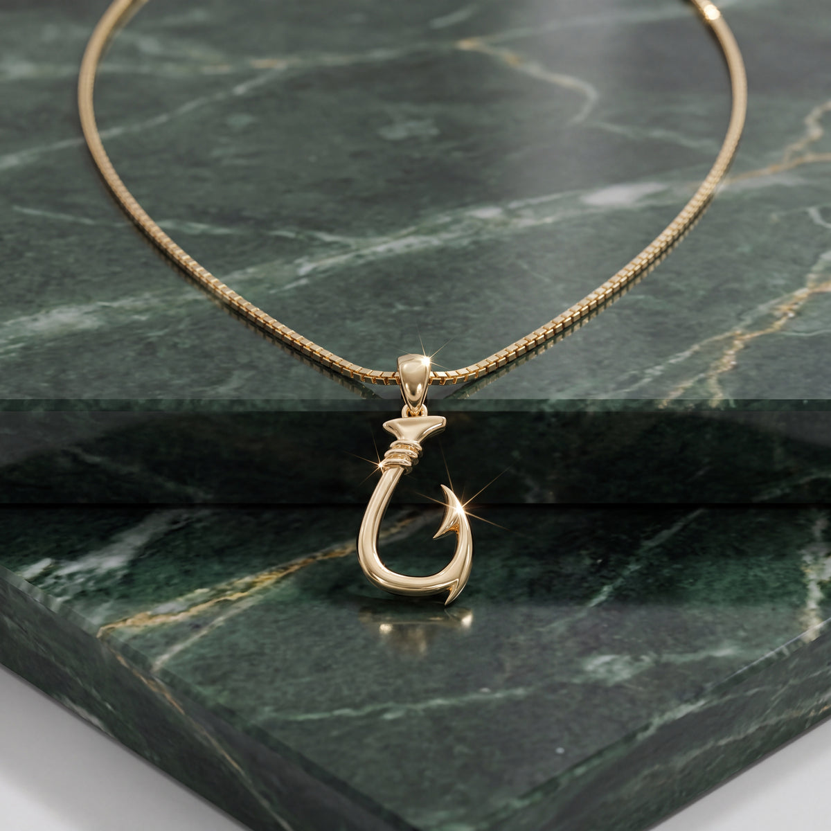 A yellow gold fish hook pendant on a box chain, resting on polished green marble.