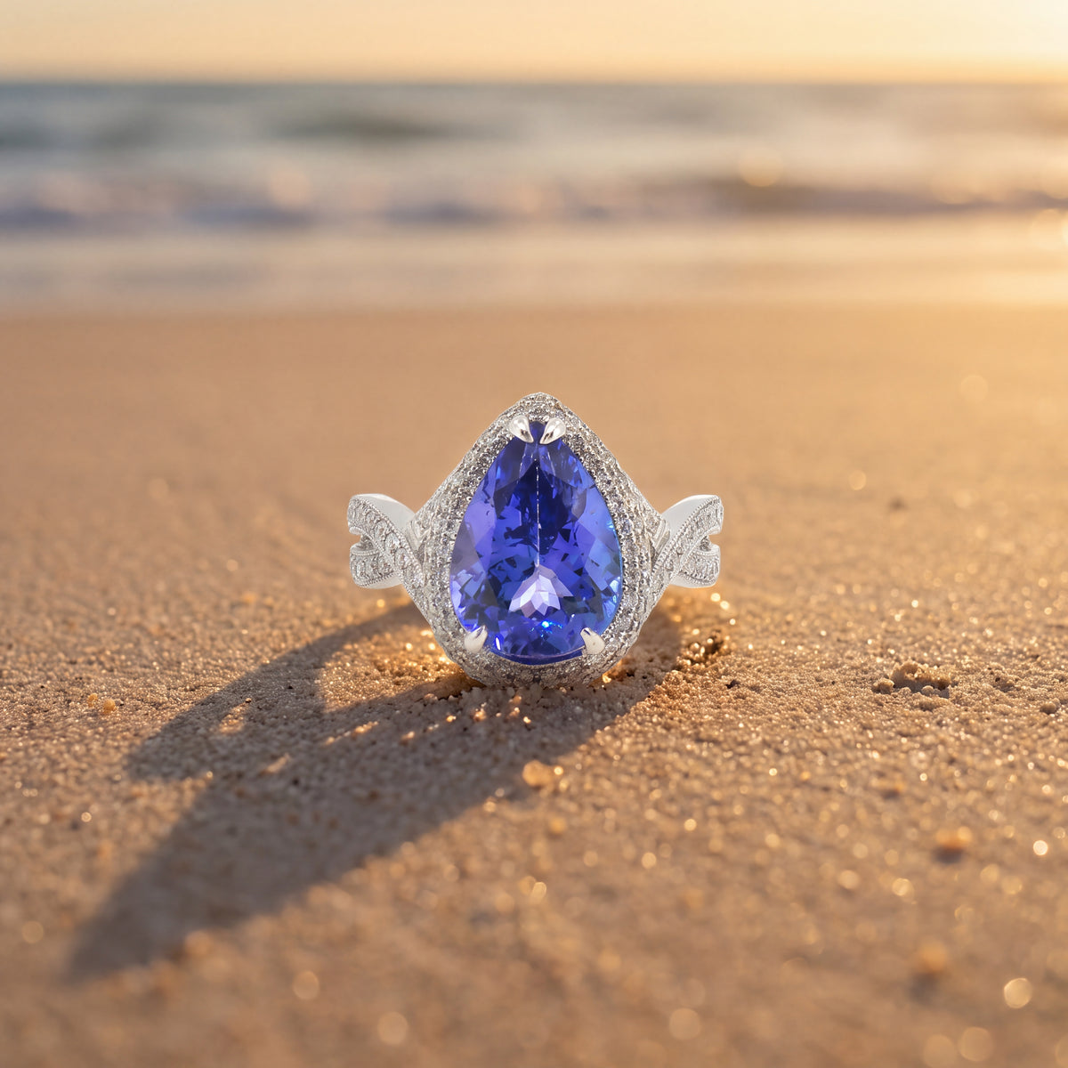 A pear-shaped violet-blue stone in a white gold halo ring, standing in sand at sunset.