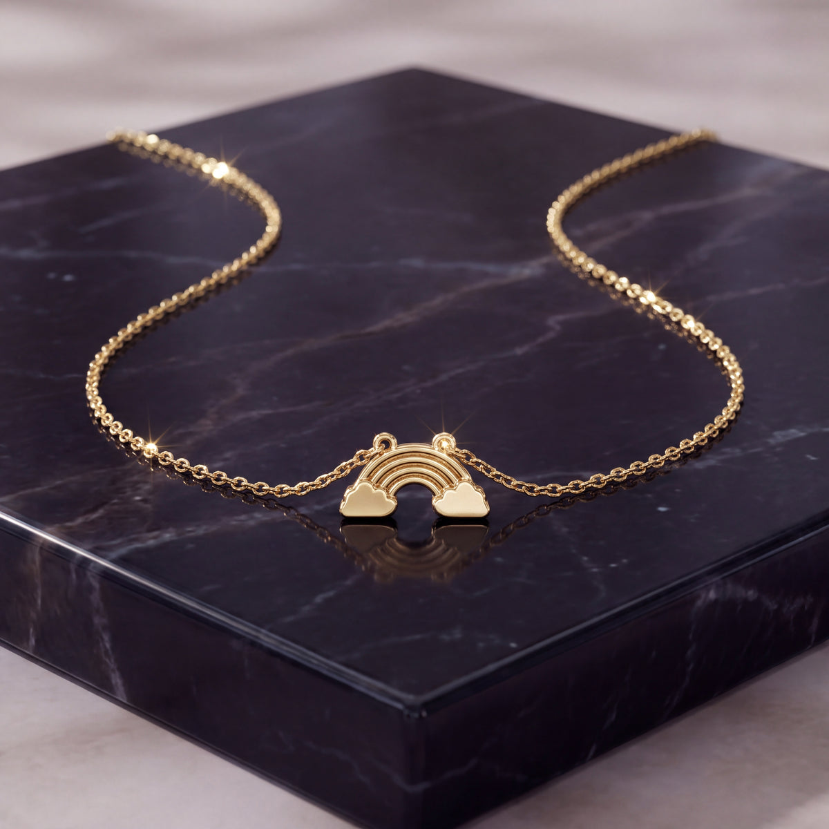 A small gold rainbow-and-cloud charm on a fine chain, laid on dark navy marble.