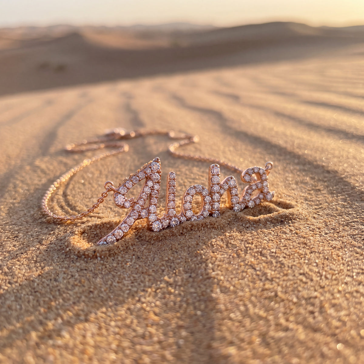 The word Aloha in diamond-set rose gold script, lying on rippled sand at sunset.