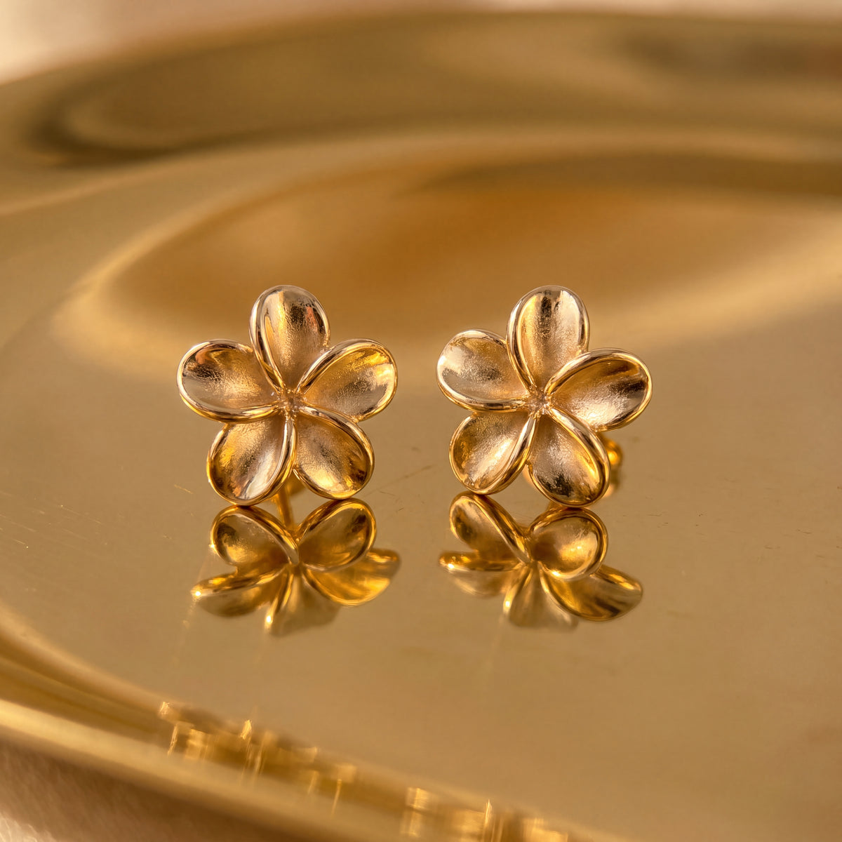 A pair of yellow gold plumeria flower stud earrings on a warm golden surface.
