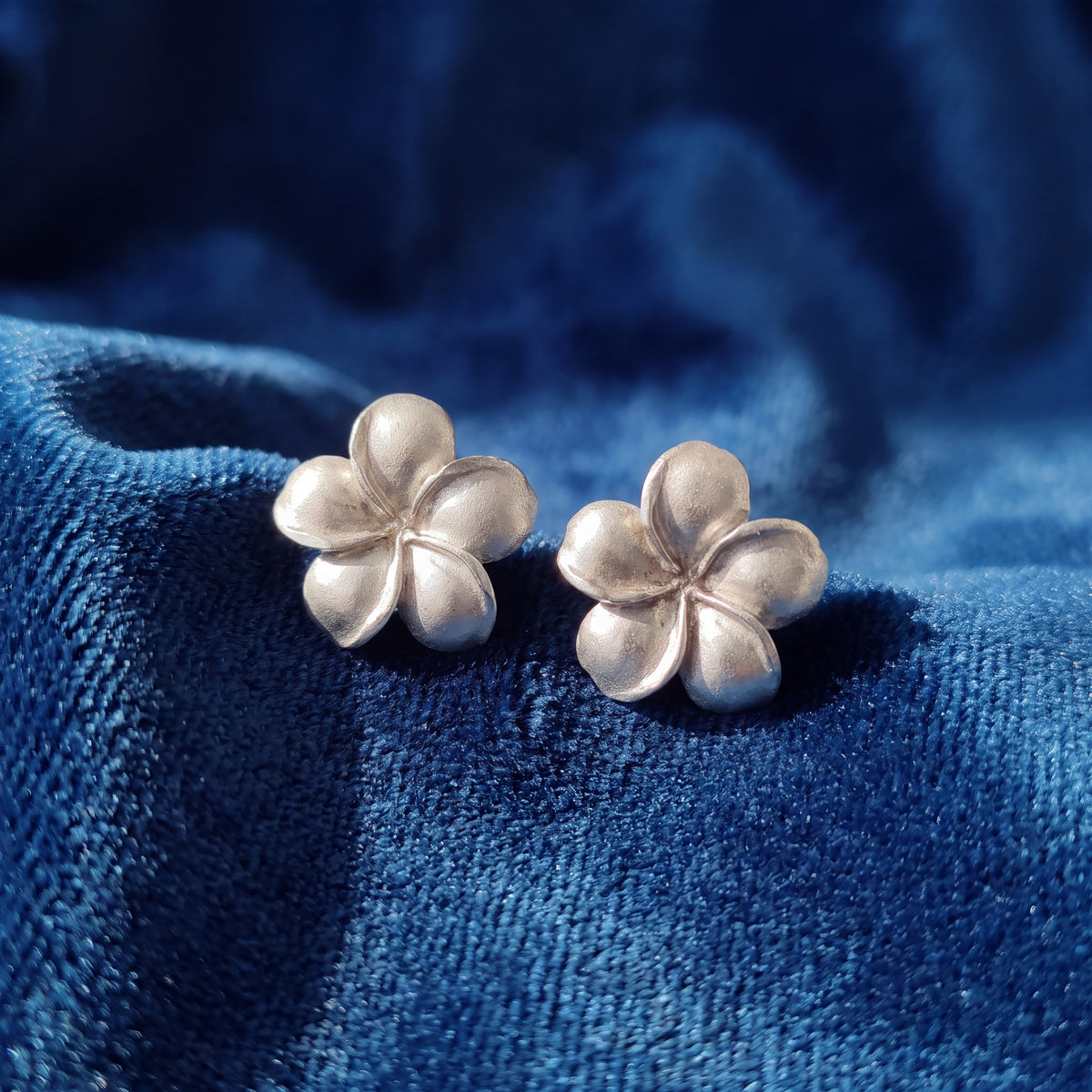 A pair of white gold plumeria flower stud earrings resting on blue velvet.
