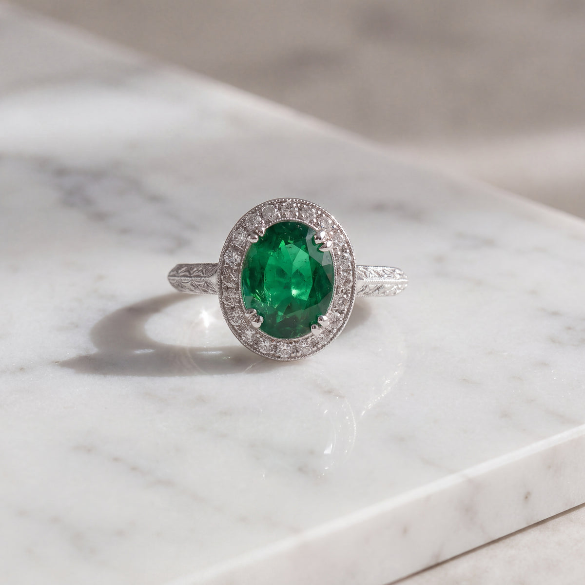Oval green emerald in four prongs with a milgrain diamond halo on an engraved white gold band