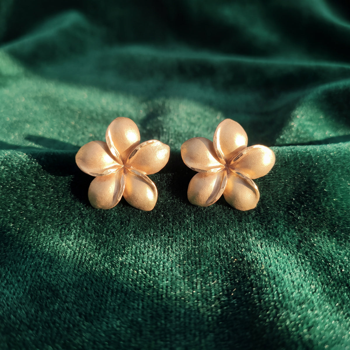 A pair of matte gold plumeria flower stud earrings resting on green velvet.