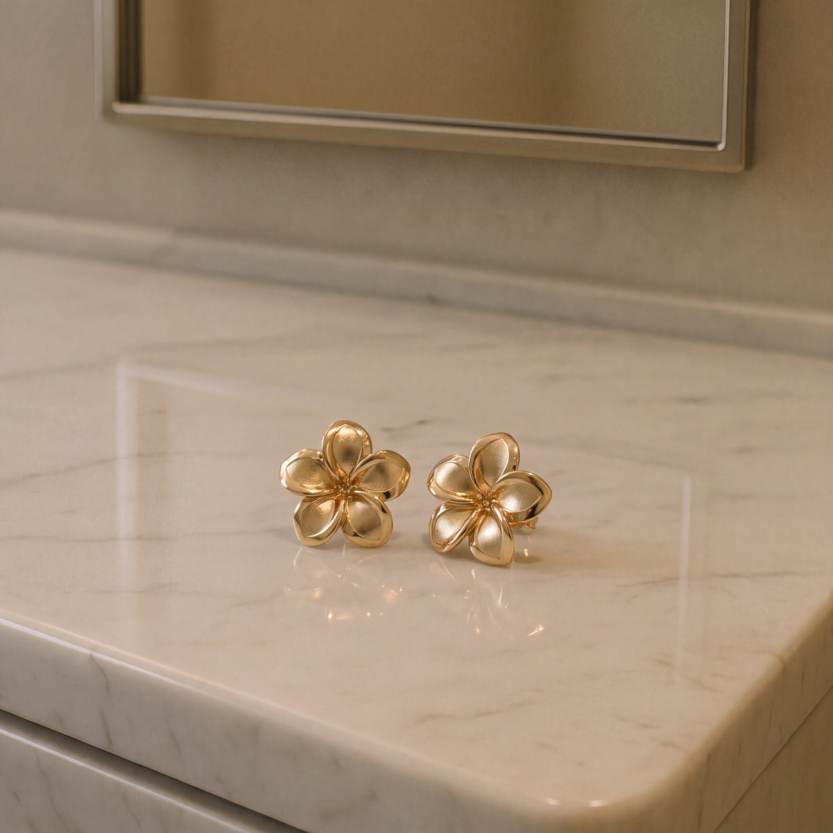 A pair of textured gold plumeria flower stud earrings on a pale marble counter.