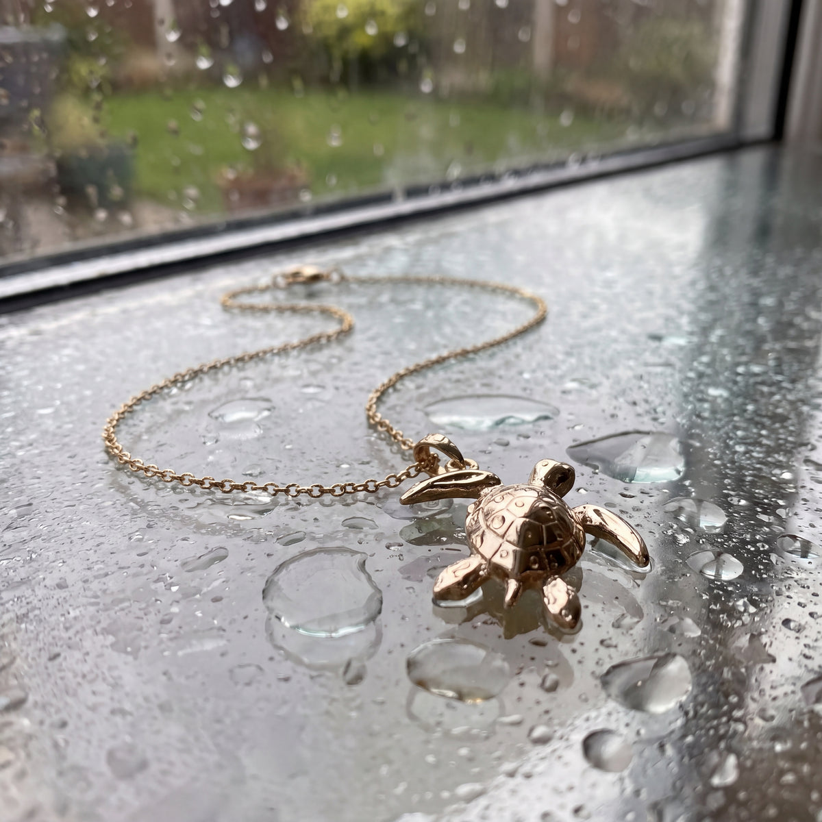 A polished gold sea turtle pendant on a fine chain, beside a rain-streaked window.