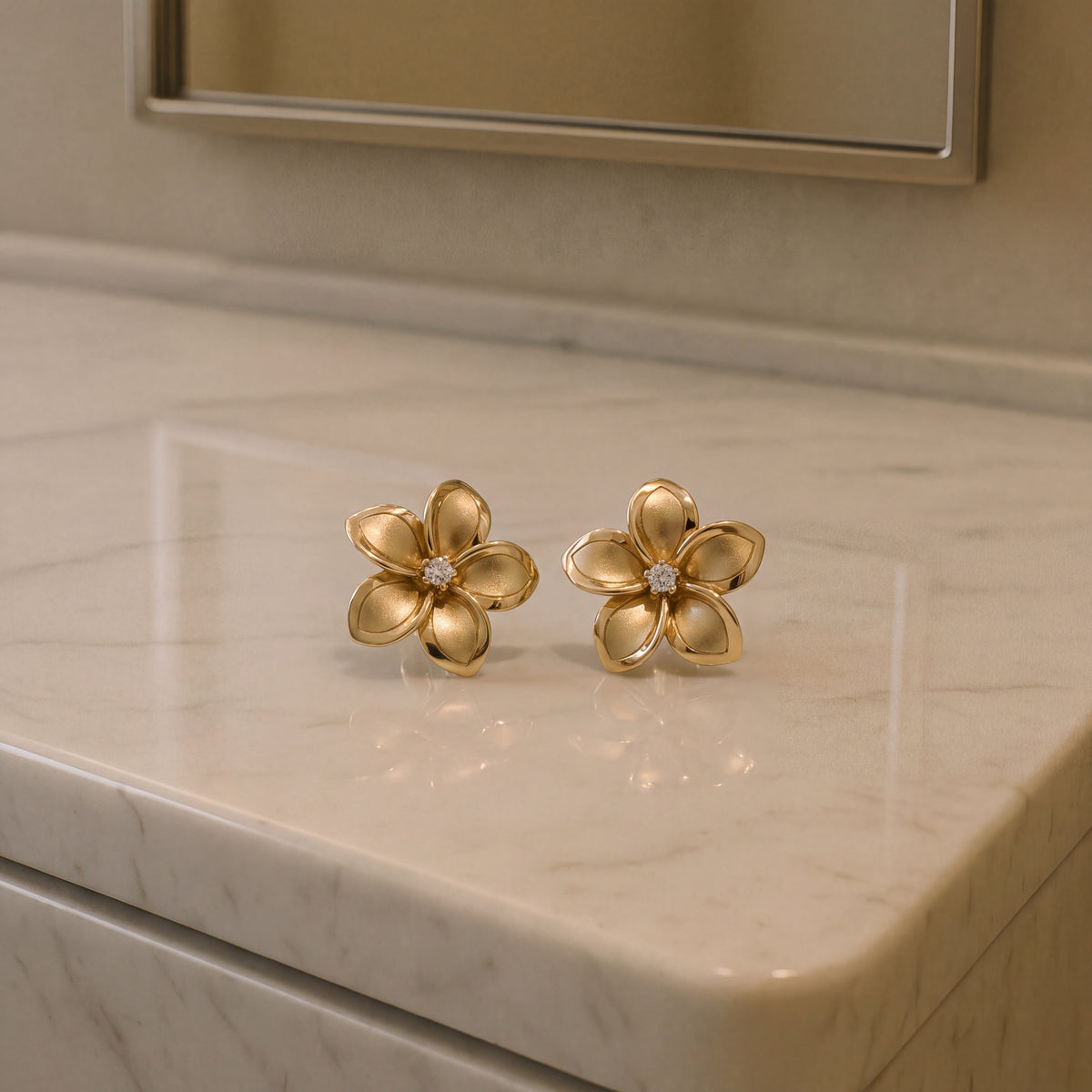 A pair of matte gold plumeria flower studs with diamond centres, on a marble counter.