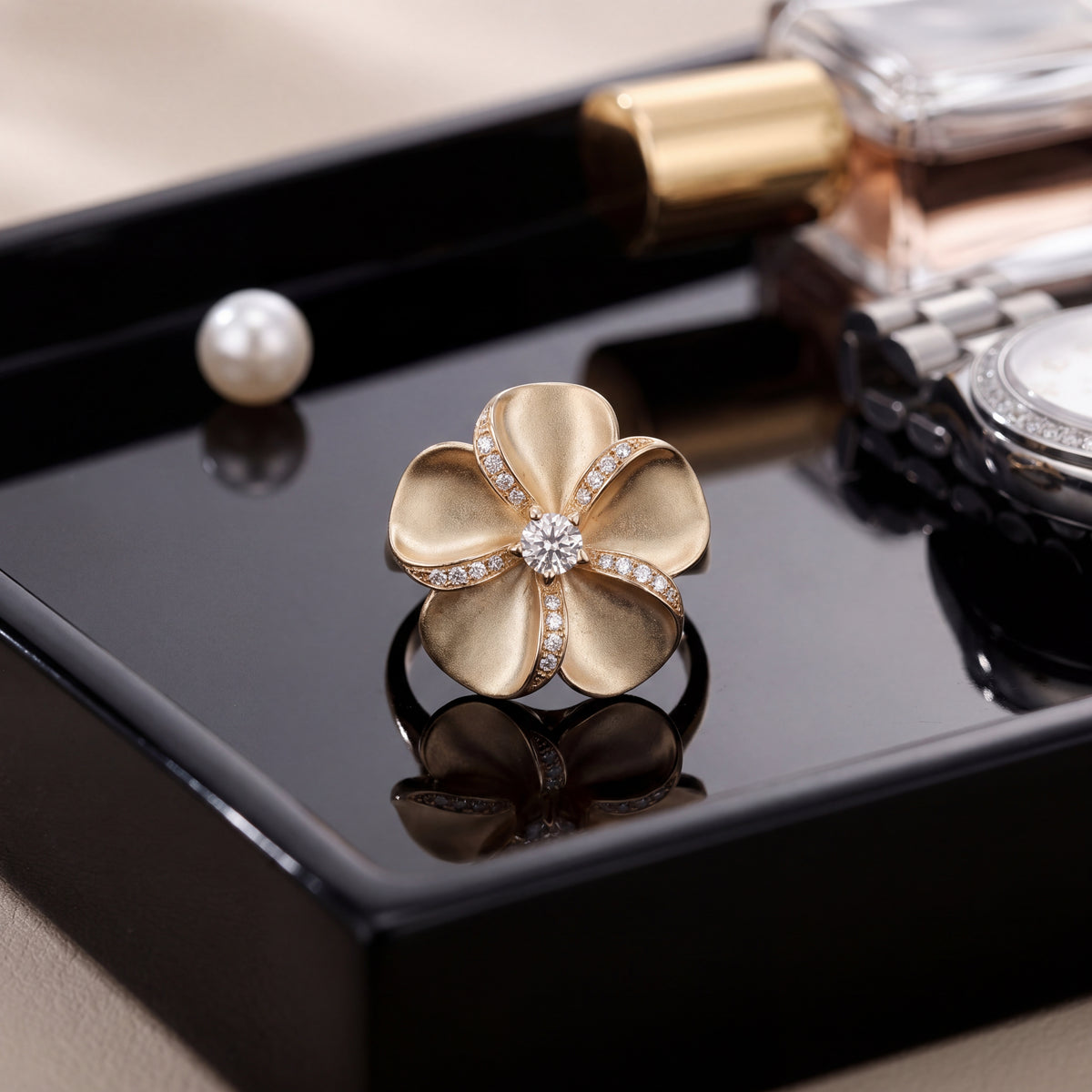 A matte gold plumeria flower cocktail ring with a diamond centre, on a black tray.