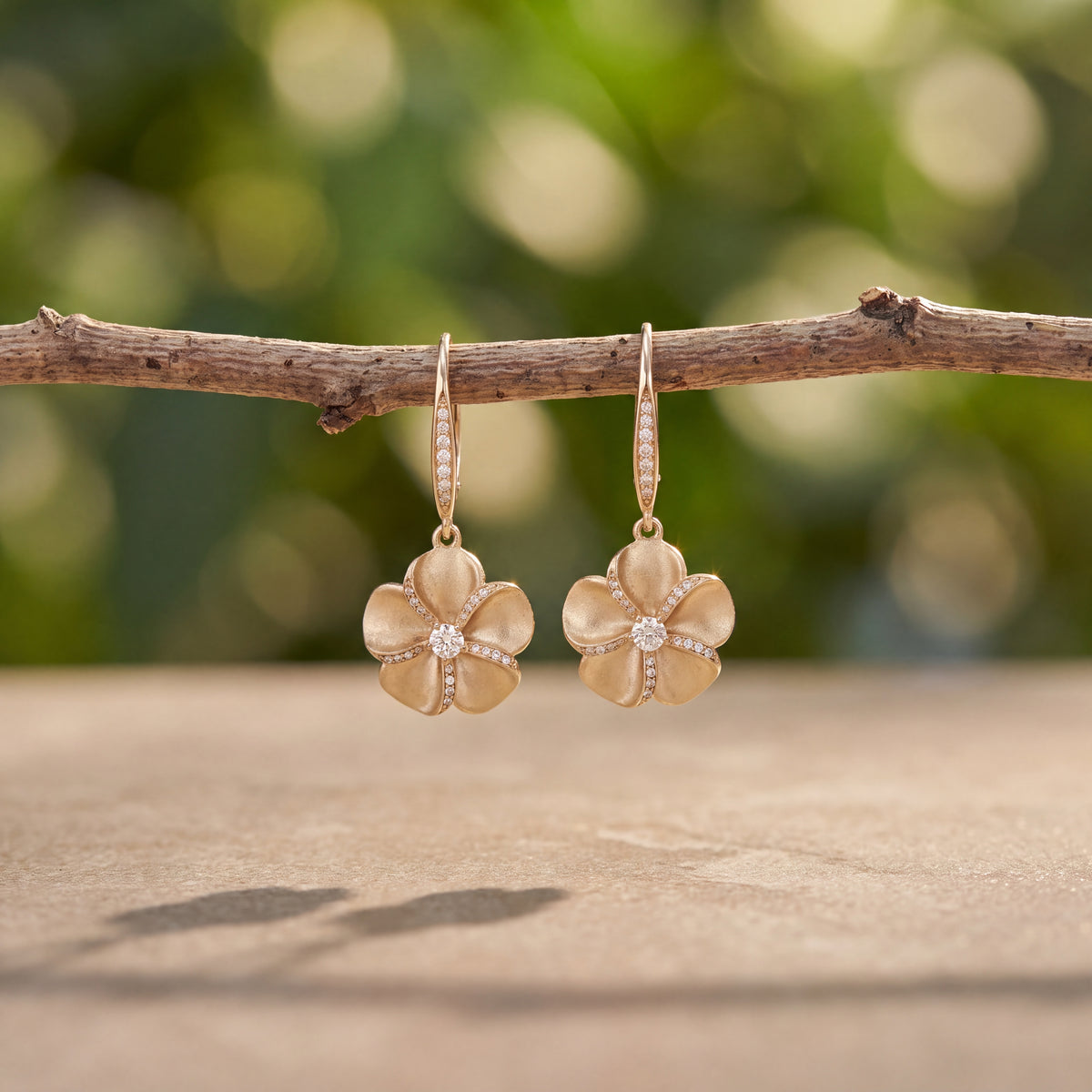 A pair of matte gold plumeria drop earrings hanging from a bare branch.