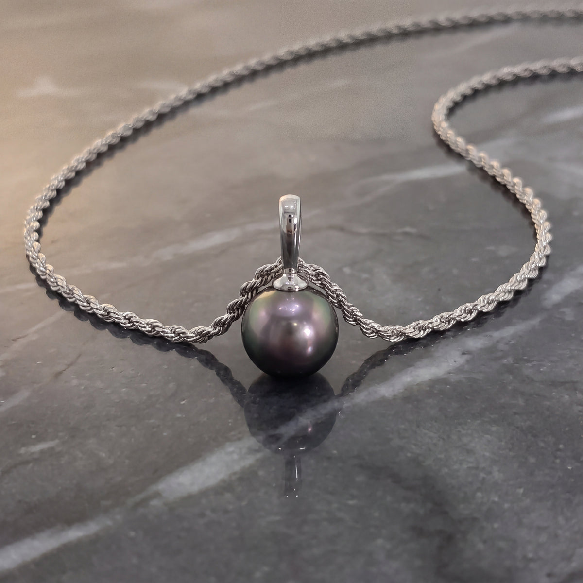 A dark grey pearl hanging from a white gold rope chain, on a grey stone surface.