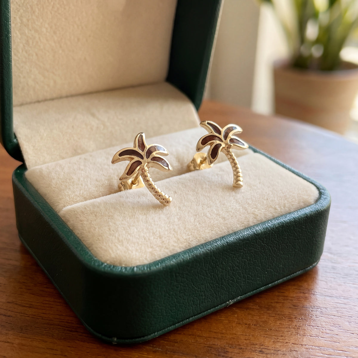A pair of gold palm tree stud earrings in an open green presentation box.