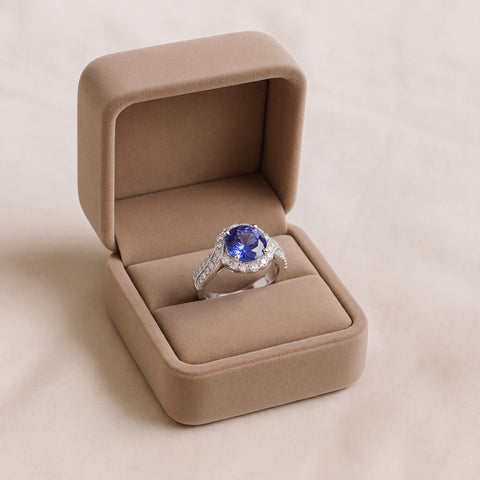 A white gold cocktail ring set with a round violet-blue tanzanite in a diamond halo, shown in an open suede box.