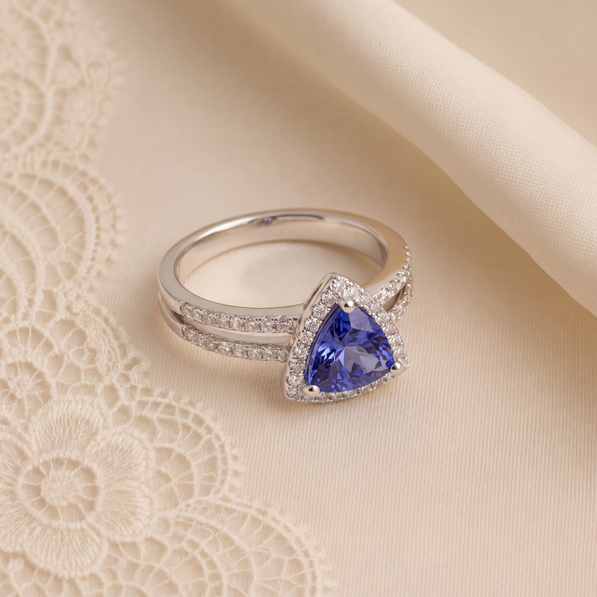 A triangular violet-blue stone in a white gold halo ring, resting on cream lace.