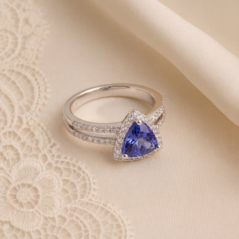 A triangular violet-blue stone in a white gold halo ring, resting on cream lace.