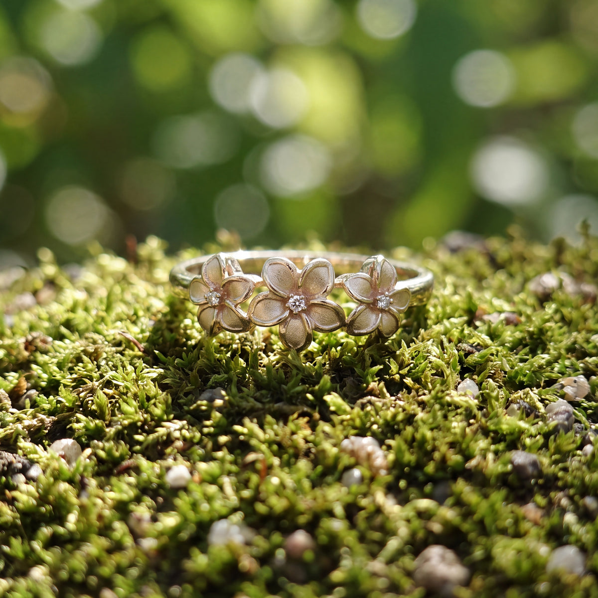 A gold ring carrying three plumeria flowers with diamond centres, resting on moss.