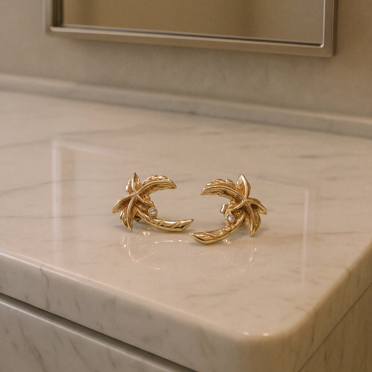 A pair of gold palm tree stud earrings with diamond accents, on a pale marble counter.