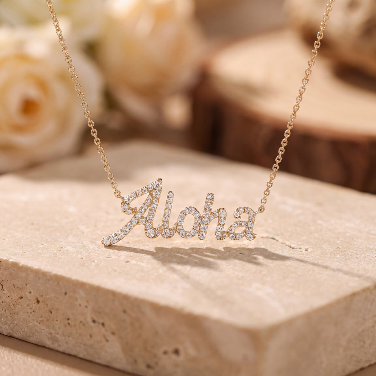 The word Aloha in diamond-set gold script, resting on pale stone beside cream roses.