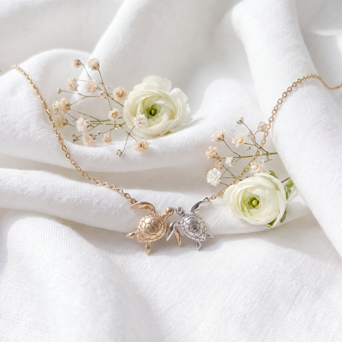 Two sea turtles side by side, one gold and one white, on a fine chain over white fabric.