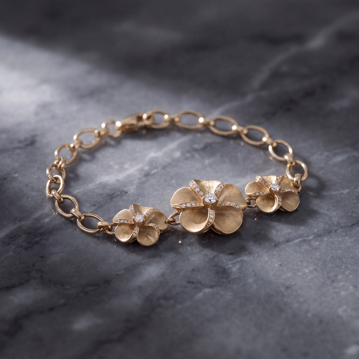 A gold chain bracelet with three plumeria flowers at its centre, on dark grey stone.