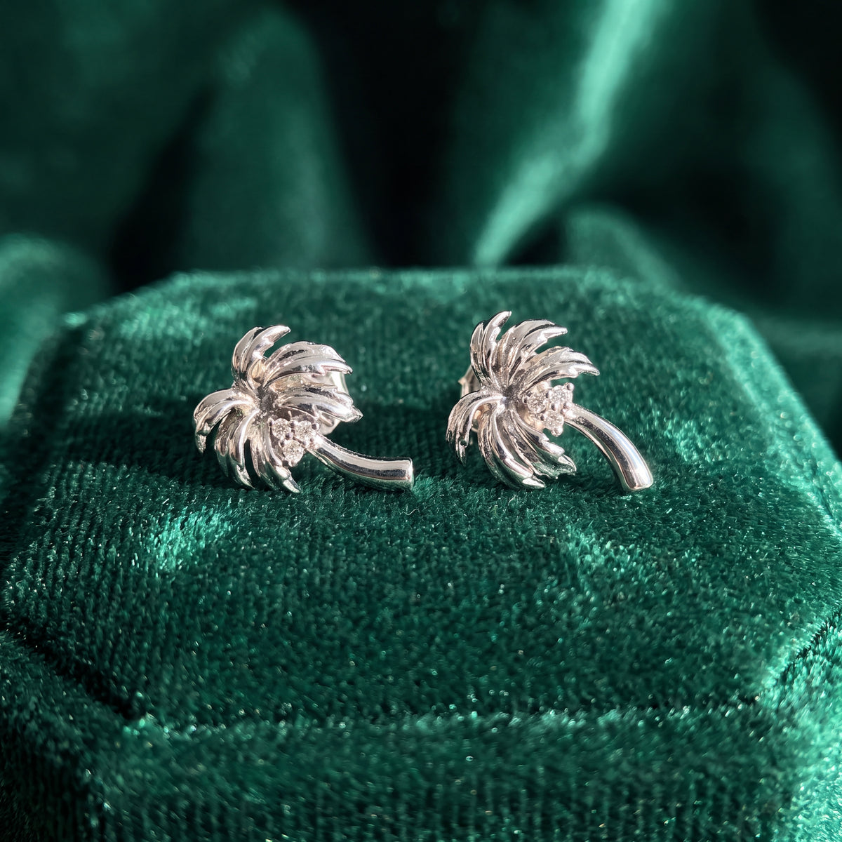 A pair of white gold palm tree stud earrings, diamonds at the centre of each swirl of fronds, resting on green velvet.