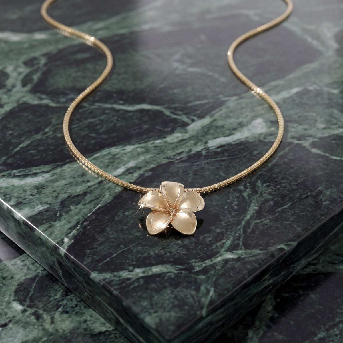 A gold plumeria flower pendant on a fine chain, resting on green marble.