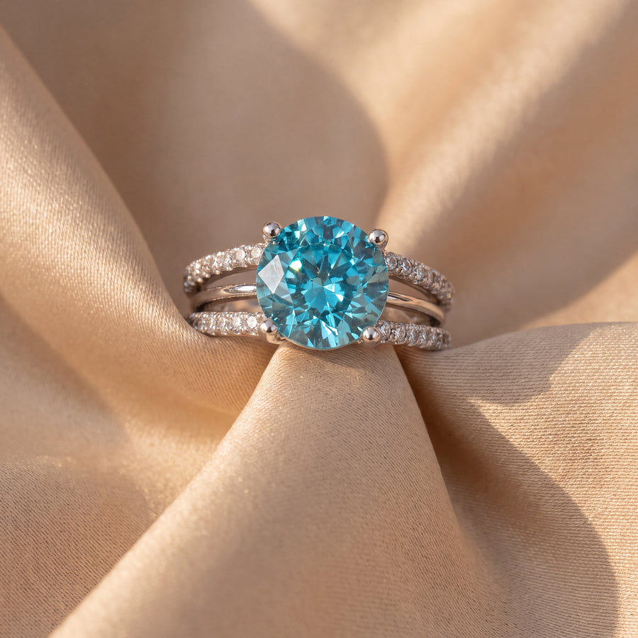 Round blue zircon in four prongs on a white gold split shank band with pavé diamonds