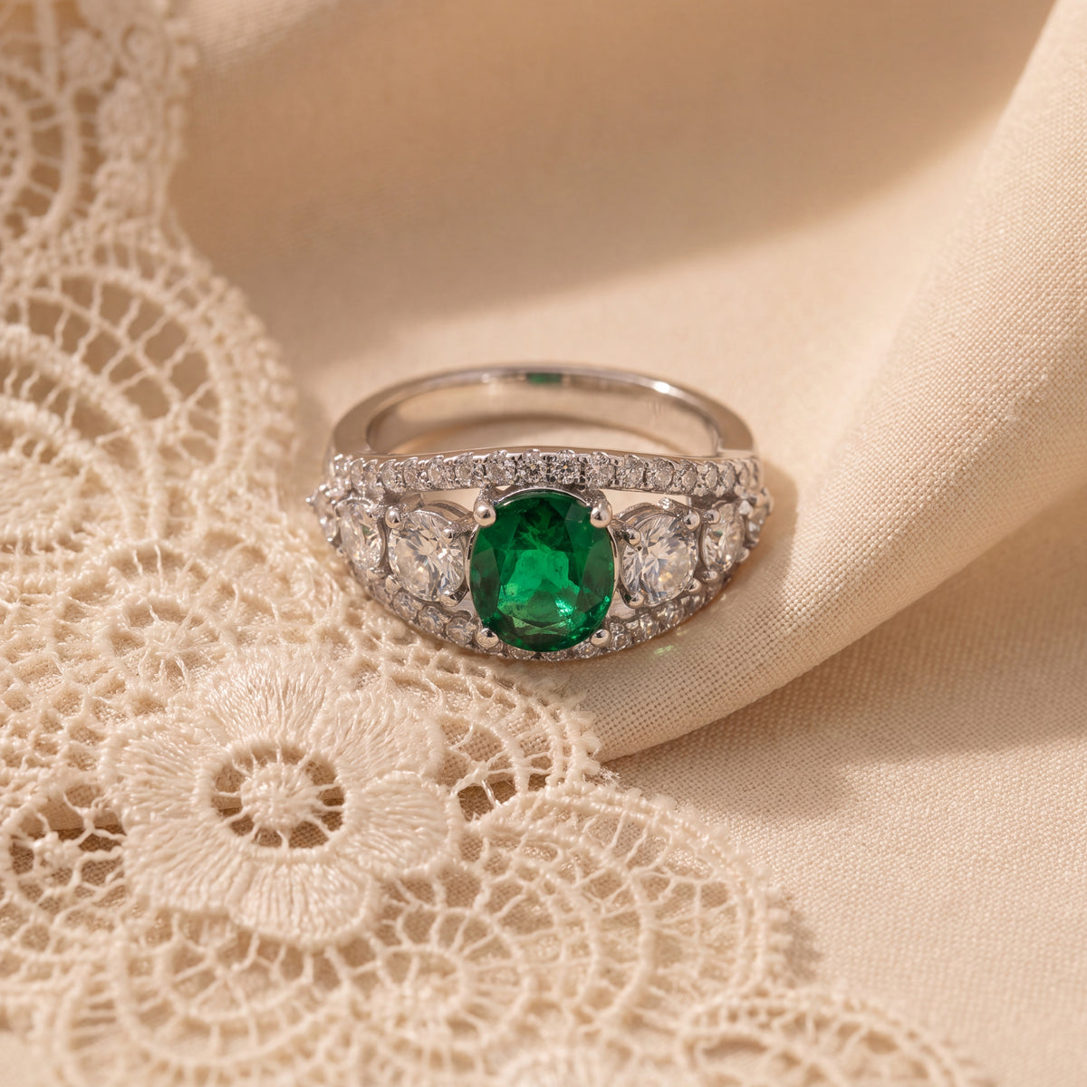 White gold ring with a green emerald center flanked by round diamonds and bordered by smaller diamonds