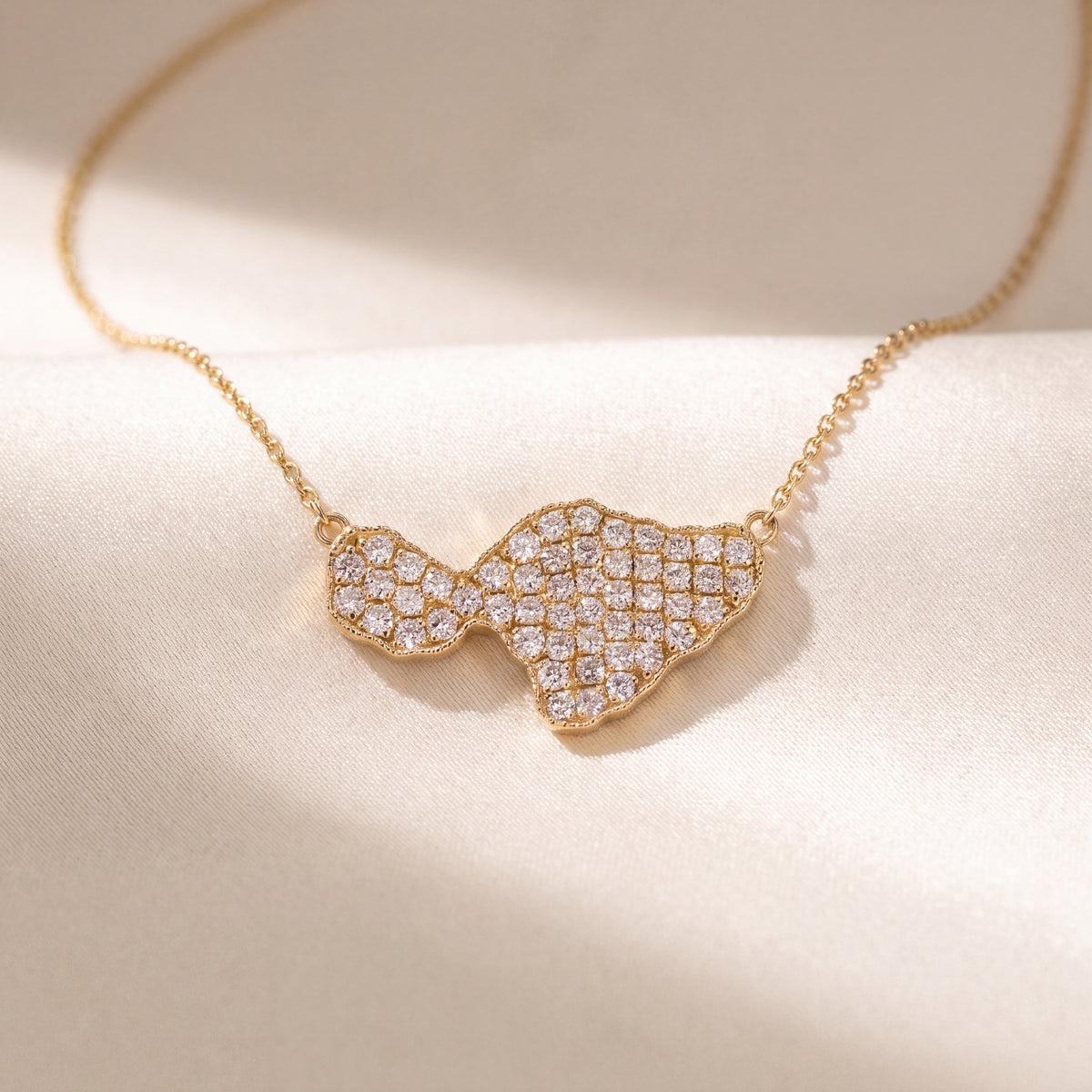 A diamond-paved island silhouette on a fine gold chain, on a pale sunlit surface.