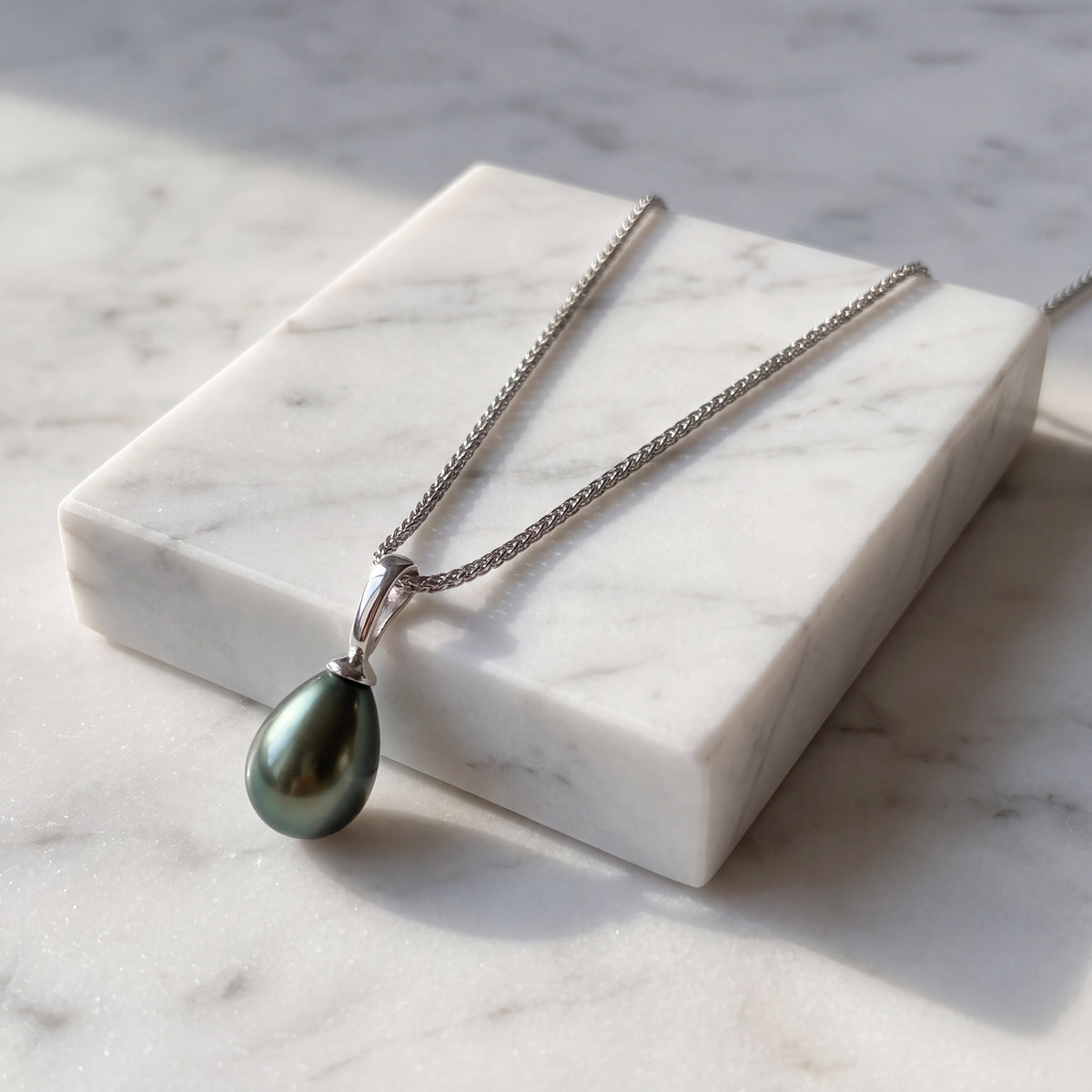 A single dark teardrop pearl on a fine white gold chain, over white marble.