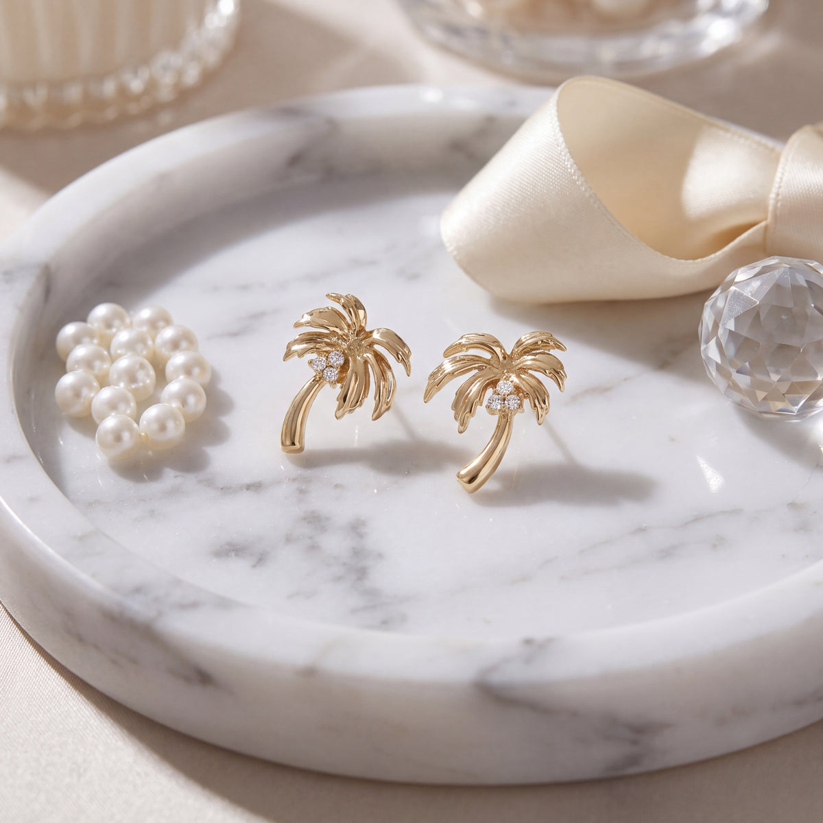 A pair of gold palm tree stud earrings on a white marble tray beside pearls and cream ribbon.
