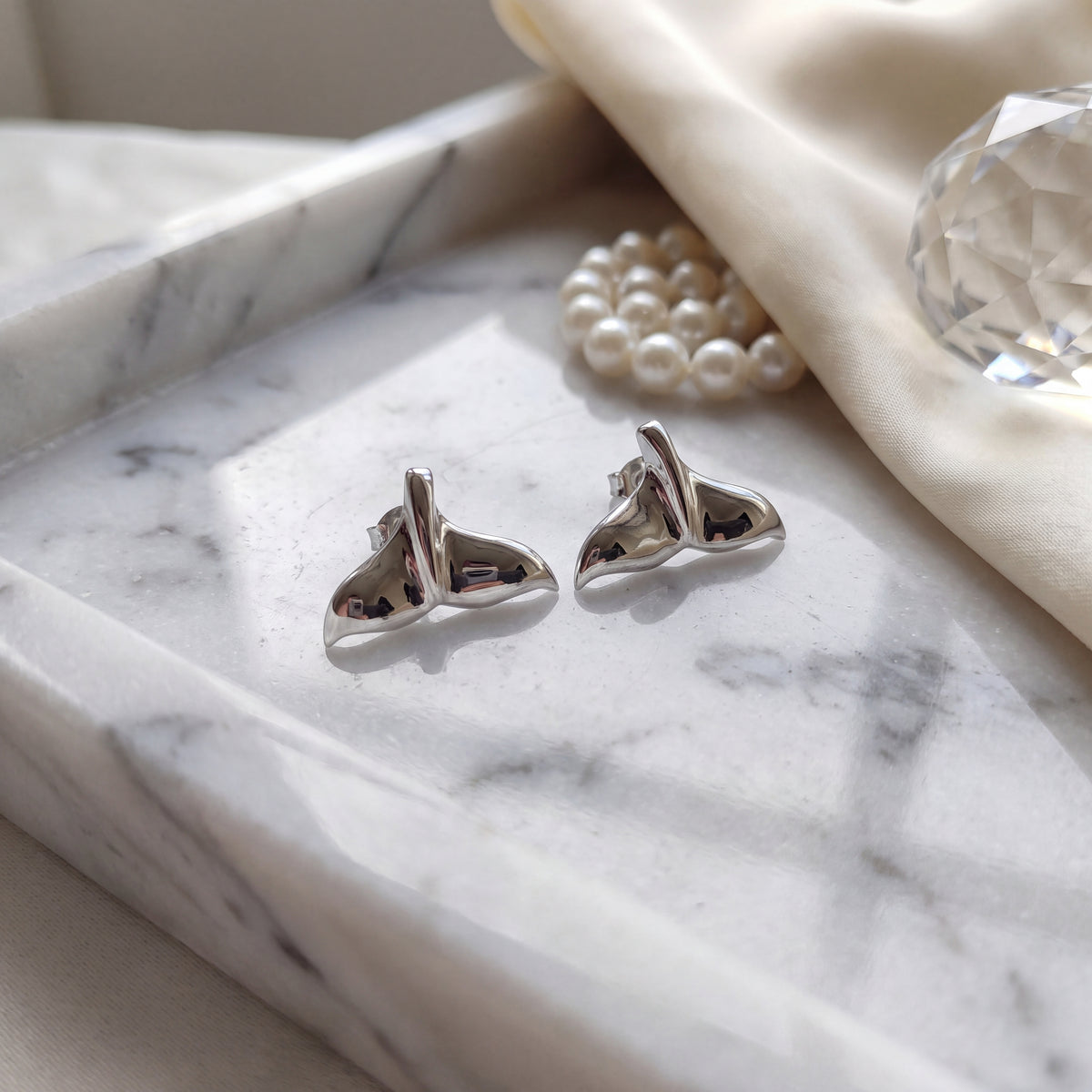A pair of polished white gold whale tail studs on marble, beside pearls and cut crystal.