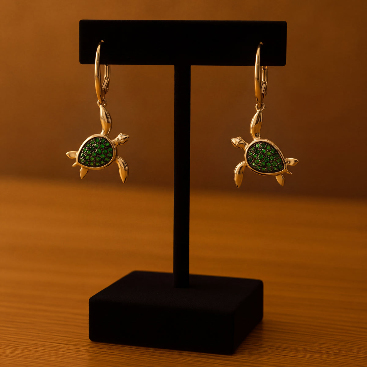 A pair of gold sea turtle drop earrings with green-set shells, on a black display stand.
