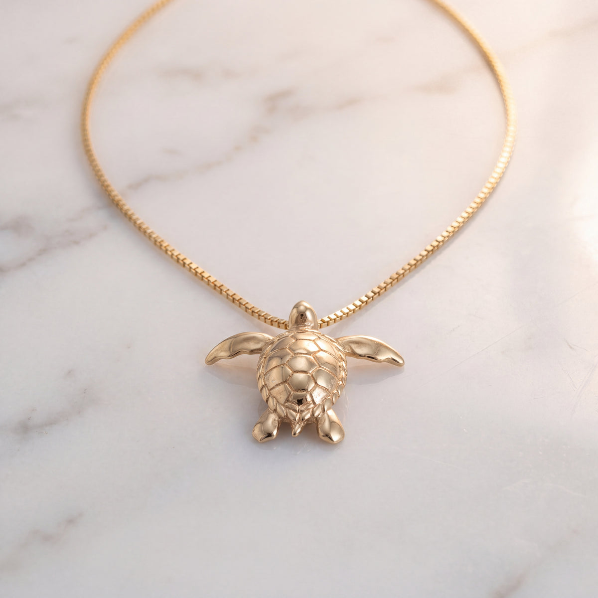 A yellow gold sea turtle charm on a fine chain, lying on white marble.
