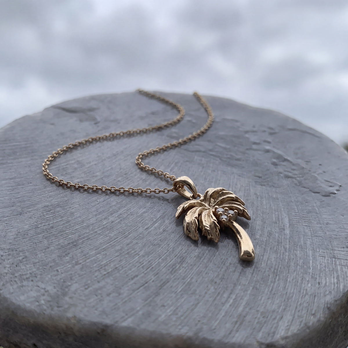 A gold palm tree pendant with diamond accents, on a fine chain over grey stone.