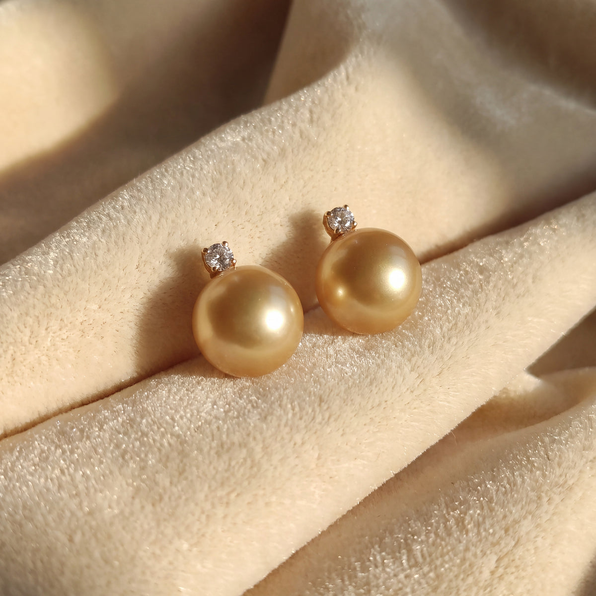 A pair of golden pearl stud earrings topped with small stones, resting on cream fabric.