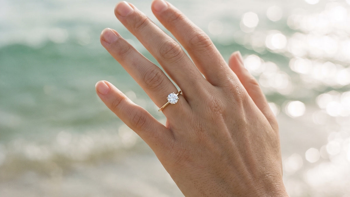 Hand wearing a solitaire diamond ring, raised against glittering ocean light